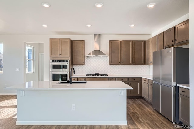 kitchen featuring a sink, light countertops, stainless steel appliances, wall chimney exhaust hood, and a kitchen island with sink