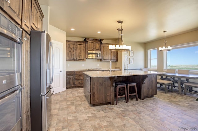 kitchen featuring appliances with stainless steel finishes, a breakfast bar area, hanging light fixtures, a kitchen island with sink, and light stone countertops