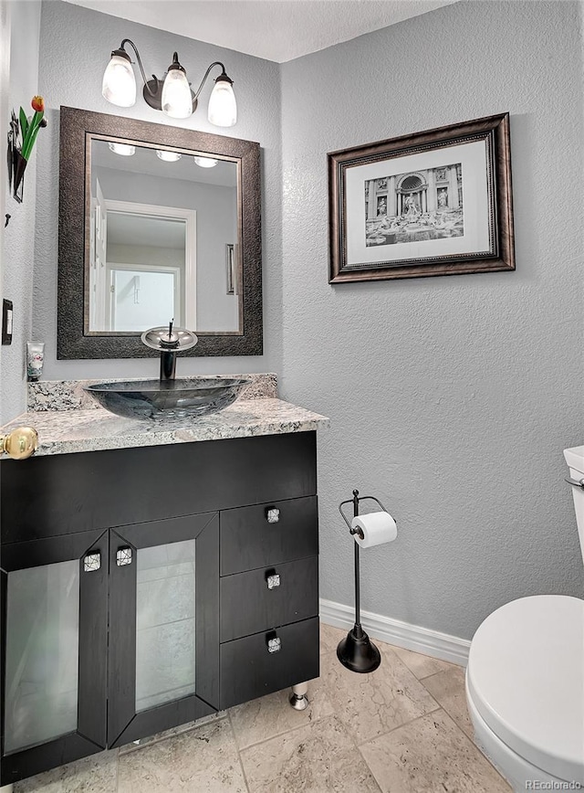 bathroom with stone tile floors, baseboards, a textured wall, toilet, and vanity