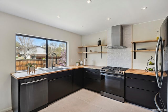 kitchen featuring open shelves, wall chimney range hood, dark cabinetry, a sink, and appliances with stainless steel finishes