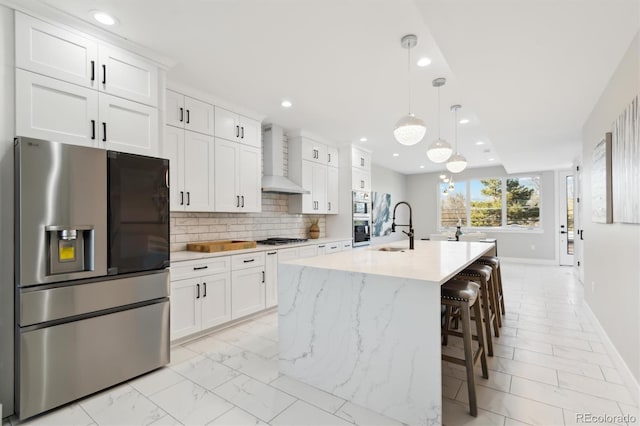 kitchen with wall chimney exhaust hood, white cabinetry, hanging light fixtures, stainless steel appliances, and a kitchen island with sink