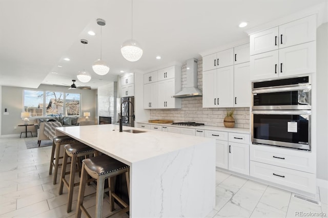 kitchen featuring sink, white cabinetry, pendant lighting, a kitchen island with sink, and wall chimney range hood