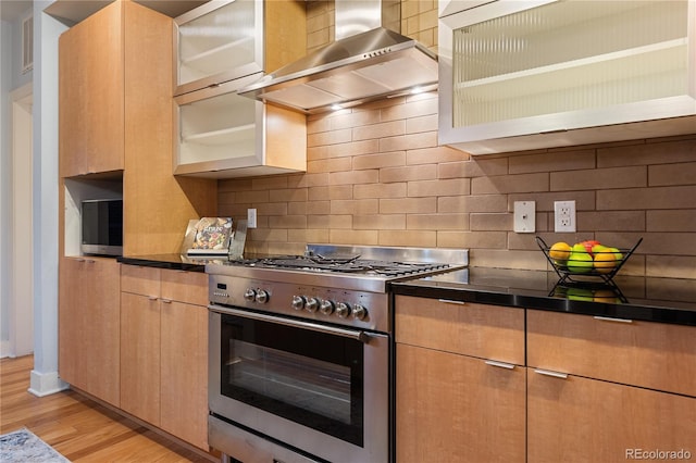 kitchen with stainless steel range, backsplash, dark countertops, wall chimney exhaust hood, and light wood finished floors