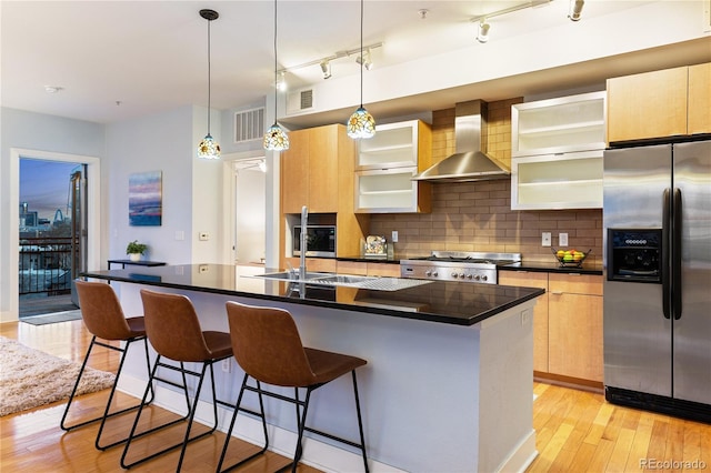 kitchen featuring light wood-type flooring, wall chimney range hood, dark countertops, stainless steel refrigerator with ice dispenser, and a kitchen island with sink