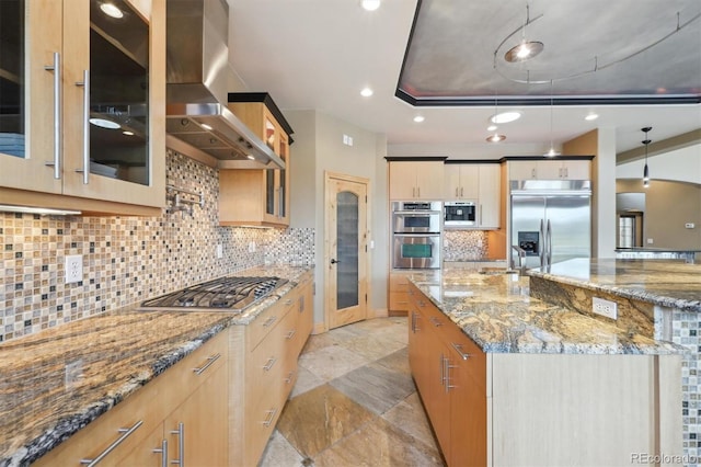 kitchen featuring light stone countertops, a tray ceiling, wall chimney range hood, built in appliances, and a large island