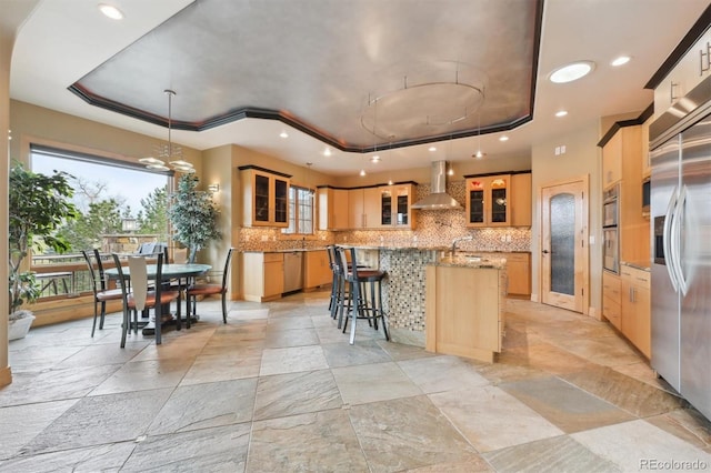 kitchen with appliances with stainless steel finishes, wall chimney exhaust hood, a raised ceiling, pendant lighting, and a center island with sink