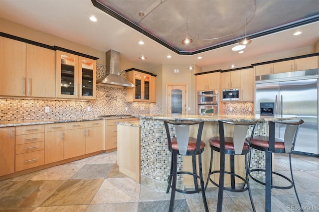 kitchen with built in appliances, wall chimney exhaust hood, light stone countertops, light brown cabinetry, and a kitchen island