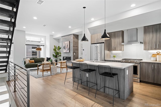 kitchen with sink, stainless steel appliances, wall chimney range hood, an island with sink, and light wood-type flooring