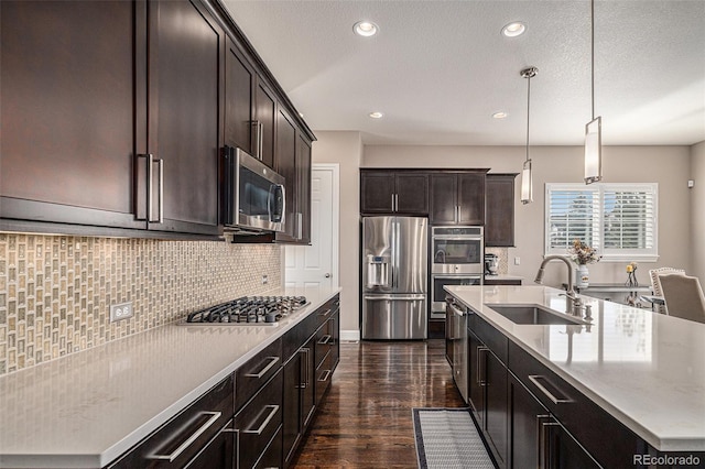 kitchen featuring sink, backsplash, stainless steel appliances, an island with sink, and decorative light fixtures