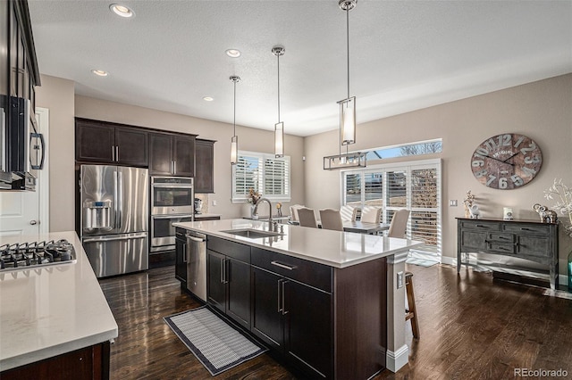 kitchen with appliances with stainless steel finishes, decorative light fixtures, an island with sink, sink, and dark wood-type flooring