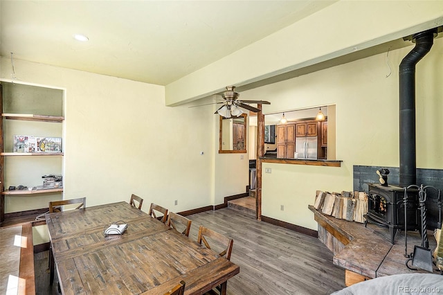 dining room with wood finished floors, baseboards, beam ceiling, a wood stove, and ceiling fan