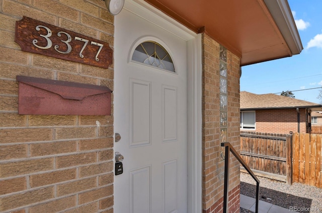 view of exterior entry with brick siding, a shingled roof, and fence