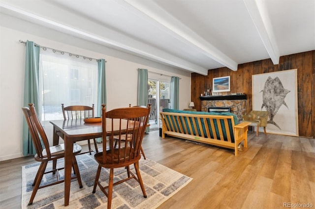 dining space featuring beam ceiling, a stone fireplace, wooden walls, and light wood-type flooring