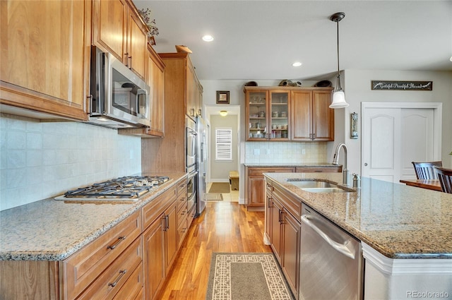 kitchen with a sink, stainless steel appliances, light wood-style floors, brown cabinetry, and glass insert cabinets