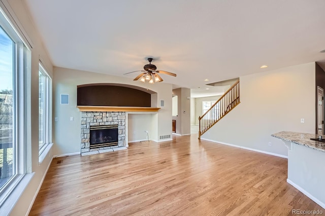 unfurnished living room with baseboards, stairs, a stone fireplace, light wood-style floors, and a ceiling fan