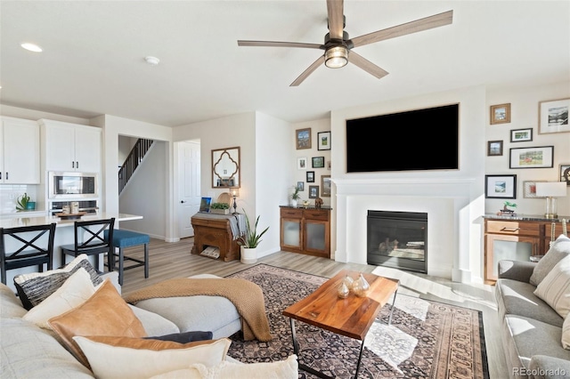 living area featuring light wood finished floors, baseboards, a ceiling fan, a glass covered fireplace, and stairs