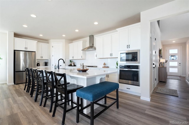 kitchen featuring stainless steel appliances, a sink, light countertops, wall chimney exhaust hood, and an island with sink