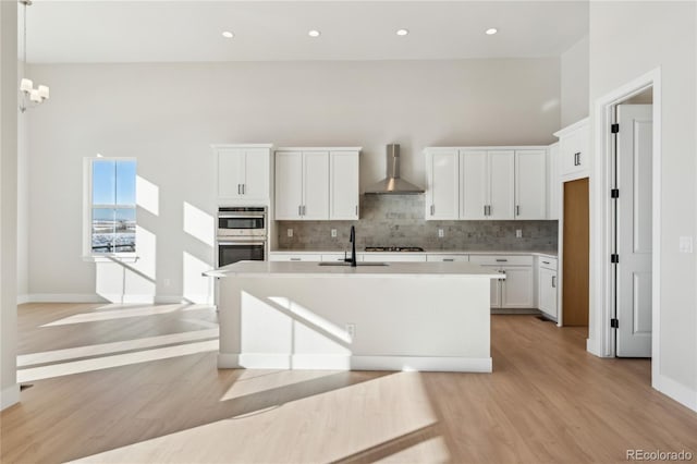 kitchen with light wood-type flooring, a kitchen island with sink, sink, wall chimney range hood, and white cabinetry