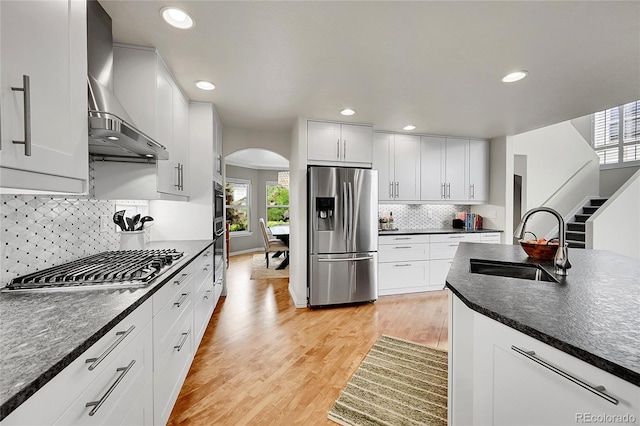 kitchen with a sink, arched walkways, wall chimney exhaust hood, stainless steel appliances, and a healthy amount of sunlight