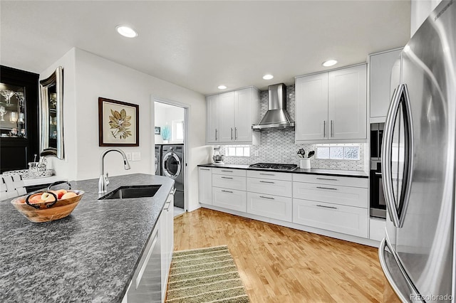 kitchen featuring wall chimney range hood, appliances with stainless steel finishes, a sink, light wood-style flooring, and tasteful backsplash