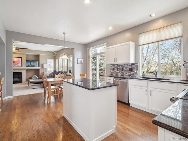 kitchen with stainless steel dishwasher, a sink, white cabinets, a glass covered fireplace, and light wood-style floors