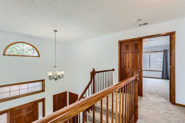 hallway with visible vents, light colored carpet, an inviting chandelier, a textured ceiling, and an upstairs landing