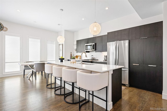 kitchen with a kitchen island with sink, a breakfast bar, light countertops, dark wood finished floors, and appliances with stainless steel finishes