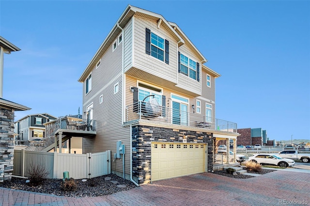 view of front facade featuring a balcony, stone siding, an attached garage, fence, and decorative driveway