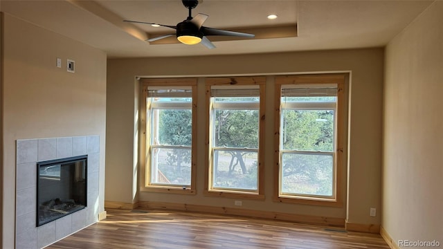 unfurnished living room with a tiled fireplace, a raised ceiling, ceiling fan, and light hardwood / wood-style flooring