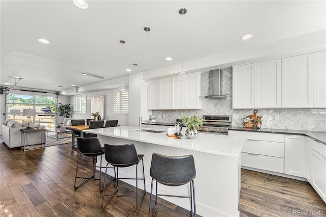 kitchen with stainless steel gas range oven, white cabinets, an island with sink, wall chimney exhaust hood, and decorative light fixtures