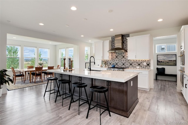 kitchen with white cabinetry, a large island, sink, wall chimney range hood, and light wood-type flooring