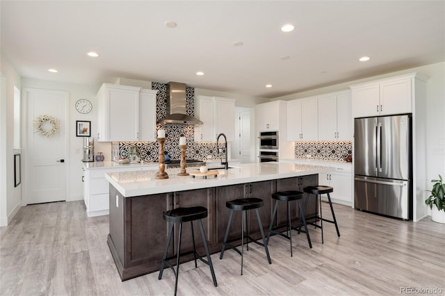 kitchen featuring sink, wall chimney exhaust hood, an island with sink, white cabinets, and appliances with stainless steel finishes