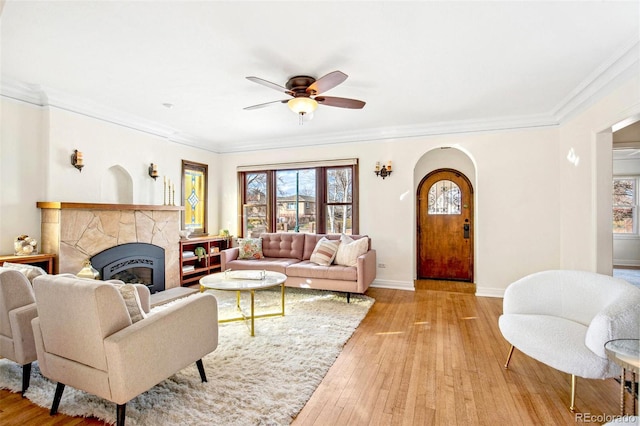 living room with arched walkways, baseboards, light wood-style flooring, ornamental molding, and a stone fireplace