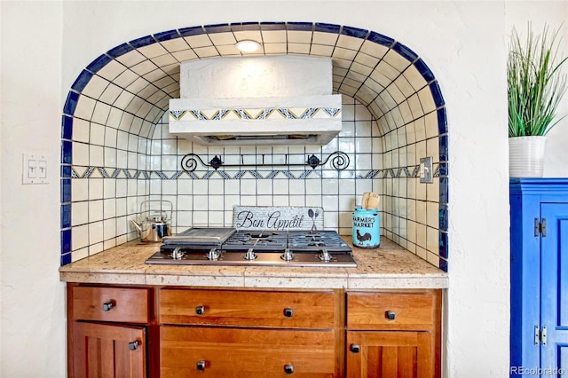 kitchen with range hood, light countertops, stainless steel gas stovetop, decorative backsplash, and brown cabinetry