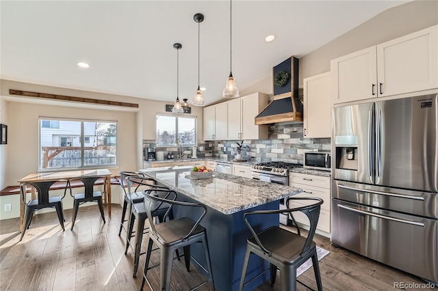 kitchen featuring wall chimney exhaust hood, stainless steel appliances, white cabinets, light stone counters, and a kitchen island