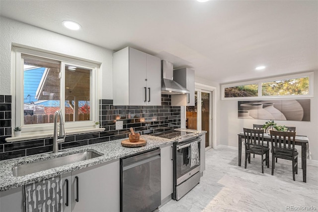 kitchen featuring appliances with stainless steel finishes, light stone counters, sink, wall chimney range hood, and backsplash