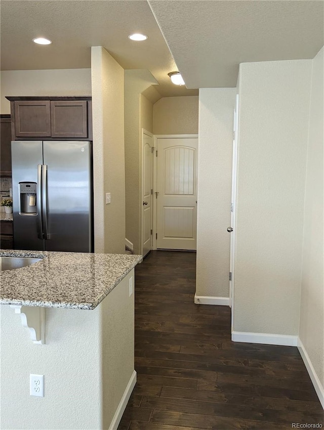 kitchen with dark wood-type flooring, stainless steel fridge, light stone countertops, dark brown cabinets, and a breakfast bar area