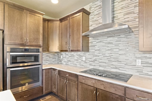 kitchen with light wood-type flooring, wall chimney exhaust hood, decorative backsplash, black cooktop, and double oven