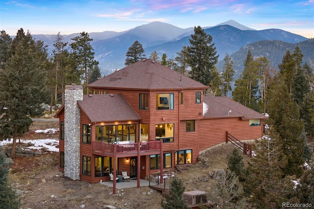 back of property at dusk featuring a shingled roof, a patio, a chimney, a deck with mountain view, and cooling unit