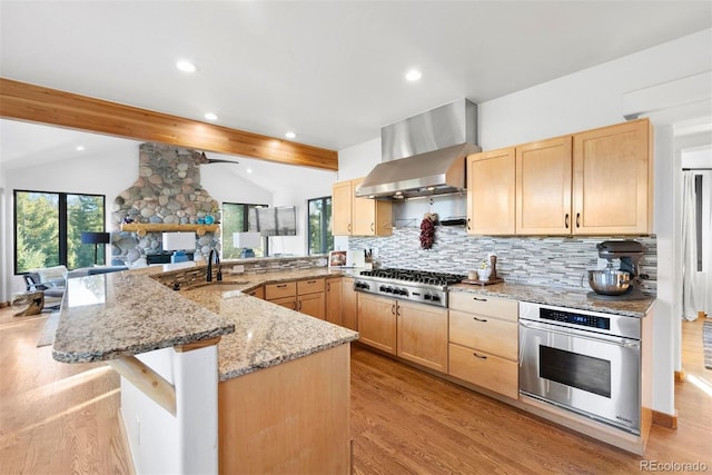 kitchen featuring appliances with stainless steel finishes, open floor plan, vaulted ceiling with beams, wall chimney range hood, and light brown cabinets