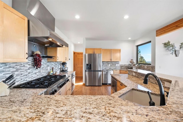 kitchen featuring stainless steel appliances, light brown cabinets, a sink, wall chimney range hood, and light stone countertops