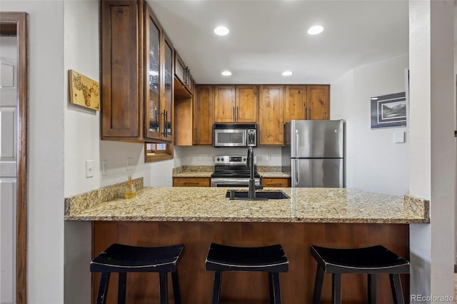 kitchen featuring glass insert cabinets, brown cabinetry, light stone counters, a peninsula, and stainless steel appliances