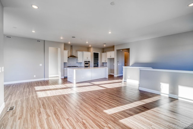 kitchen with a center island with sink, light wood-type flooring, white cabinets, wall chimney range hood, and tasteful backsplash