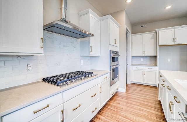 kitchen featuring appliances with stainless steel finishes, wall chimney exhaust hood, white cabinets, light hardwood / wood-style flooring, and backsplash