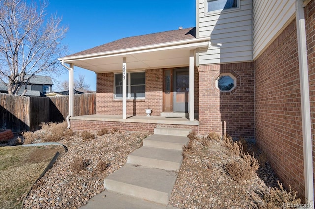 doorway to property with covered porch, brick siding, a shingled roof, and fence