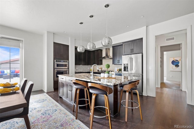 kitchen with wall chimney range hood, backsplash, a kitchen island with sink, hanging light fixtures, and appliances with stainless steel finishes