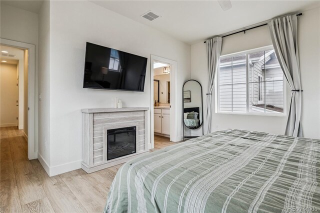 bedroom with a glass covered fireplace, light wood-type flooring, ensuite bath, baseboards, and visible vents