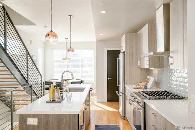 kitchen with high quality appliances, modern cabinets, an island with sink, light wood-style floors, and wall chimney range hood