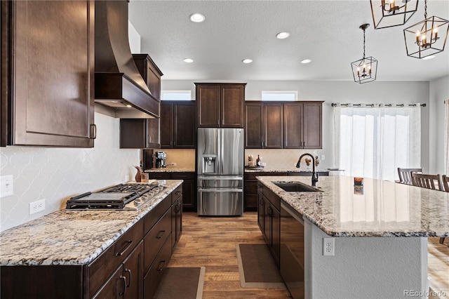 kitchen featuring sink, custom exhaust hood, pendant lighting, stainless steel appliances, and a kitchen island with sink