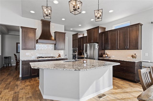 kitchen with hanging light fixtures, stainless steel appliances, custom range hood, and an island with sink